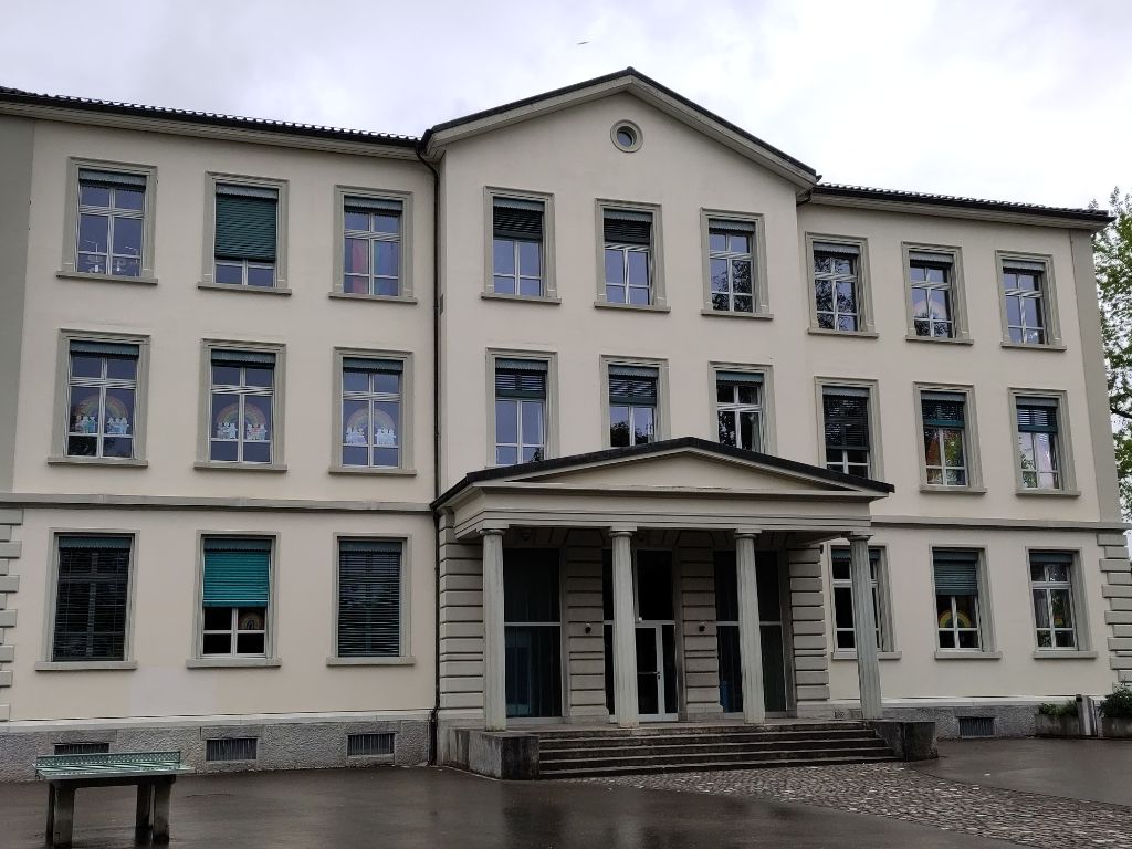 Foto des Schulhauses Obermatt. An den Fenstern aller Klassenzimmer sind Regenbogen aus Papier sichtbar.